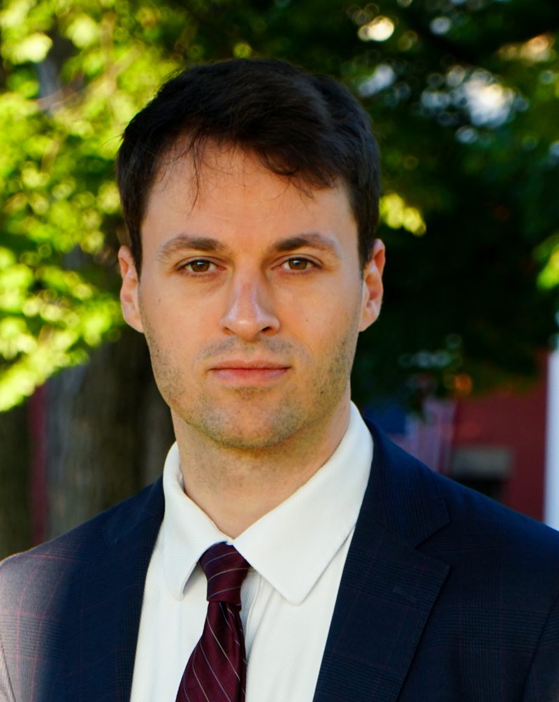 Thomas Plan, head and shoulders, in a navy suit and burgundy tie.