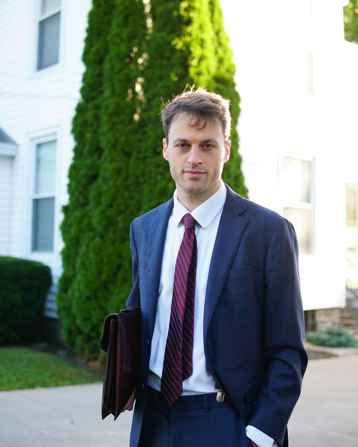 Thomas Plan in a navy suit, a briefcase under his arm, outside a white house in Penn Yan.